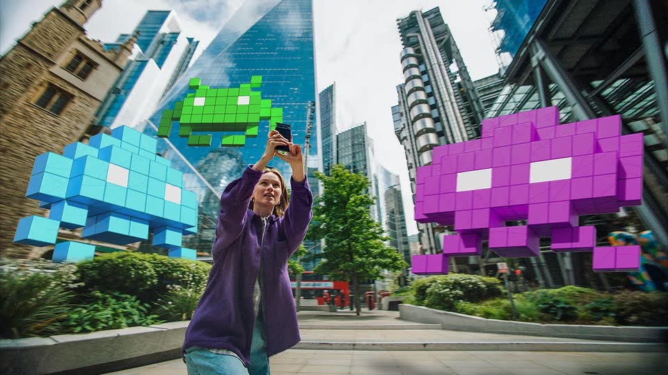 A player aiming a phone at giant pixel invaders between city towers